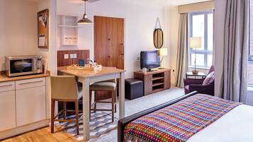 A compact hotel room with a kitchenette, a bed with a colorful blanket, a television, and a seating area with a chair and a sofa.
