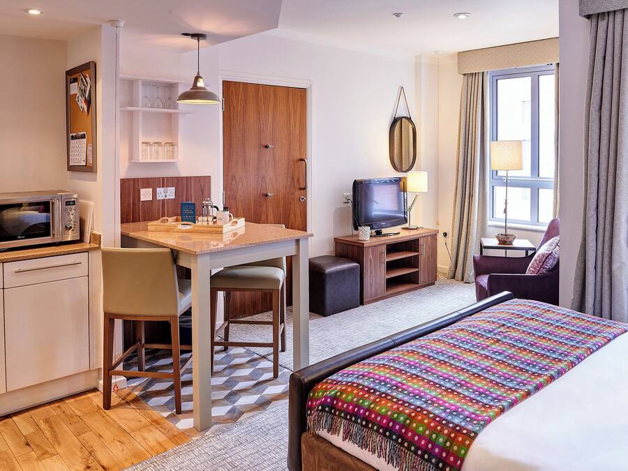 A compact hotel room with a kitchenette, a bed with a colorful blanket, a television, and a seating area with a chair and a sofa.