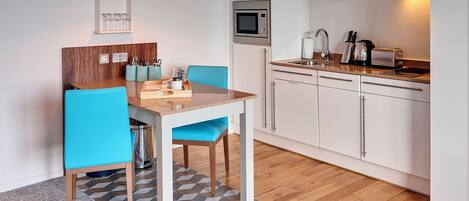 A modern kitchen with white cabinets, a microwave, and a toaster. There is a dining table with blue chairs and a wooden floor.