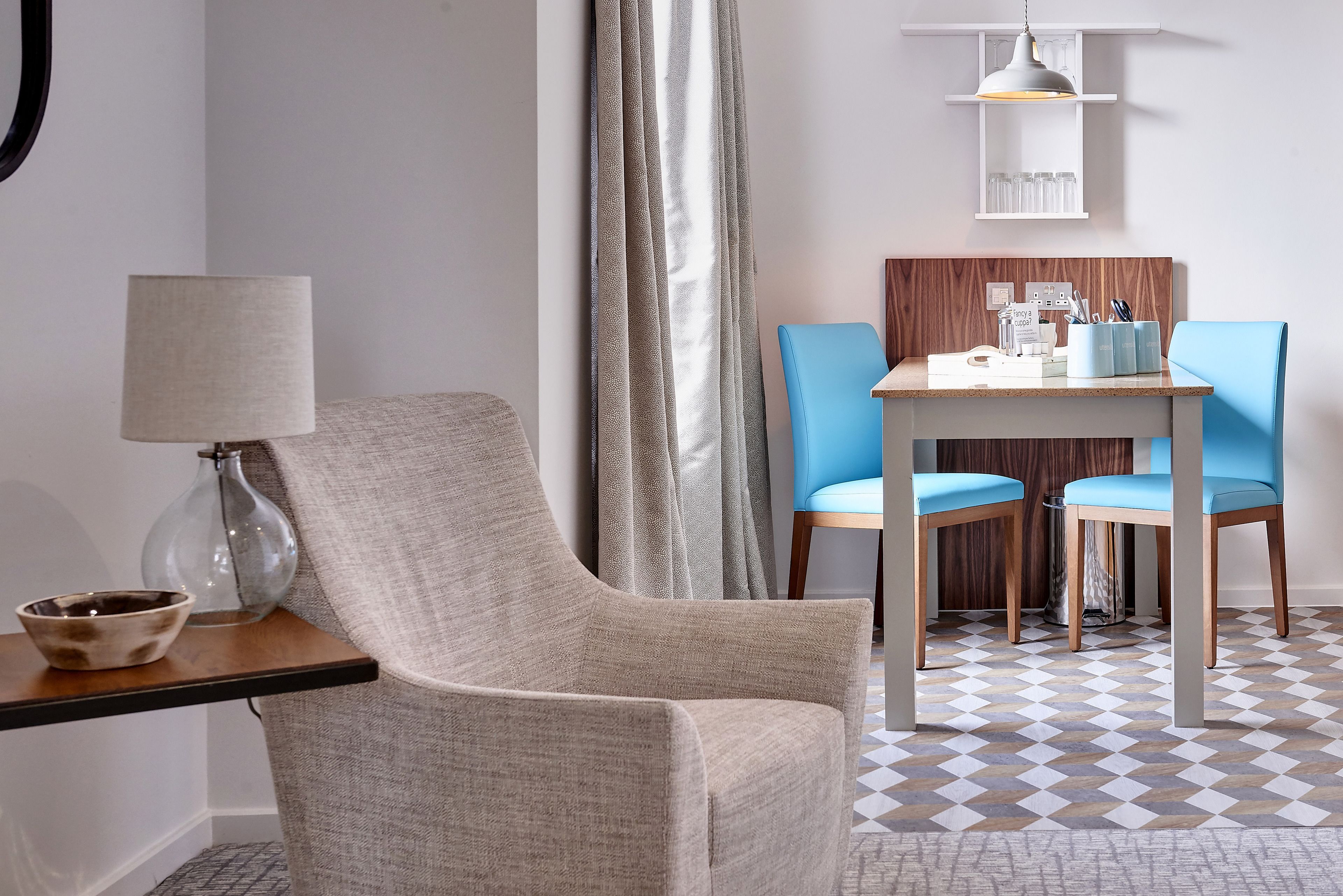 Quarto de hotel com design moderno, com uma poltrona cinza, uma mesinha com luminária, uma cadeira azul e uma mesa de jantar com painel de madeira.