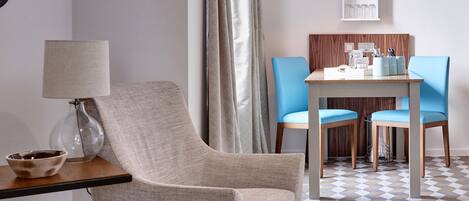 A hotel room with a modern design, featuring a grey armchair, a small table with a lamp, a blue chair, and a dining table with a wooden panel.