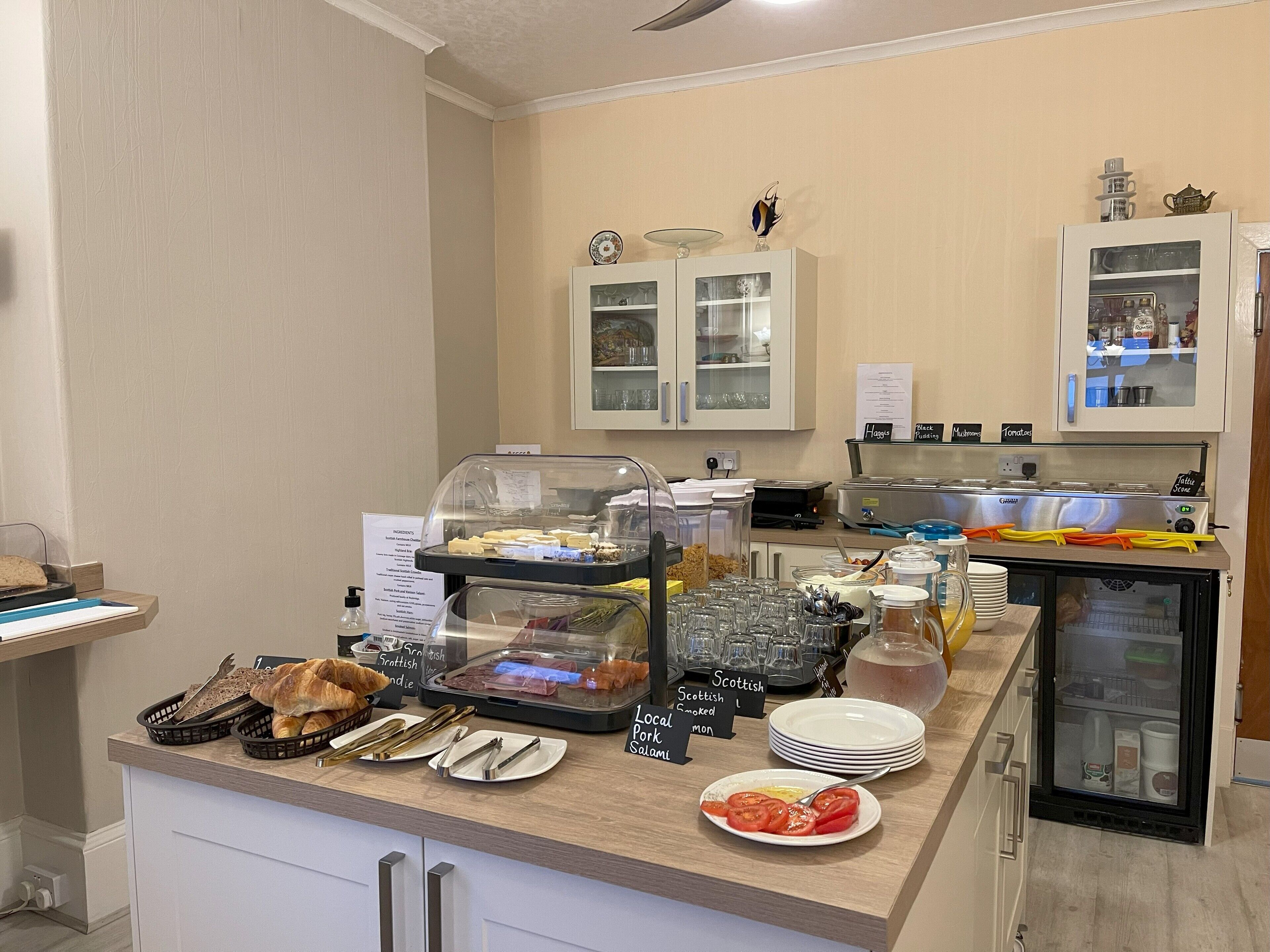 Un área de bufé con diversas opciones de comida, incluyendo un expositor de vidrio con sándwiches, una barra con platos y cubiertos, y un refrigerador con aderezos.