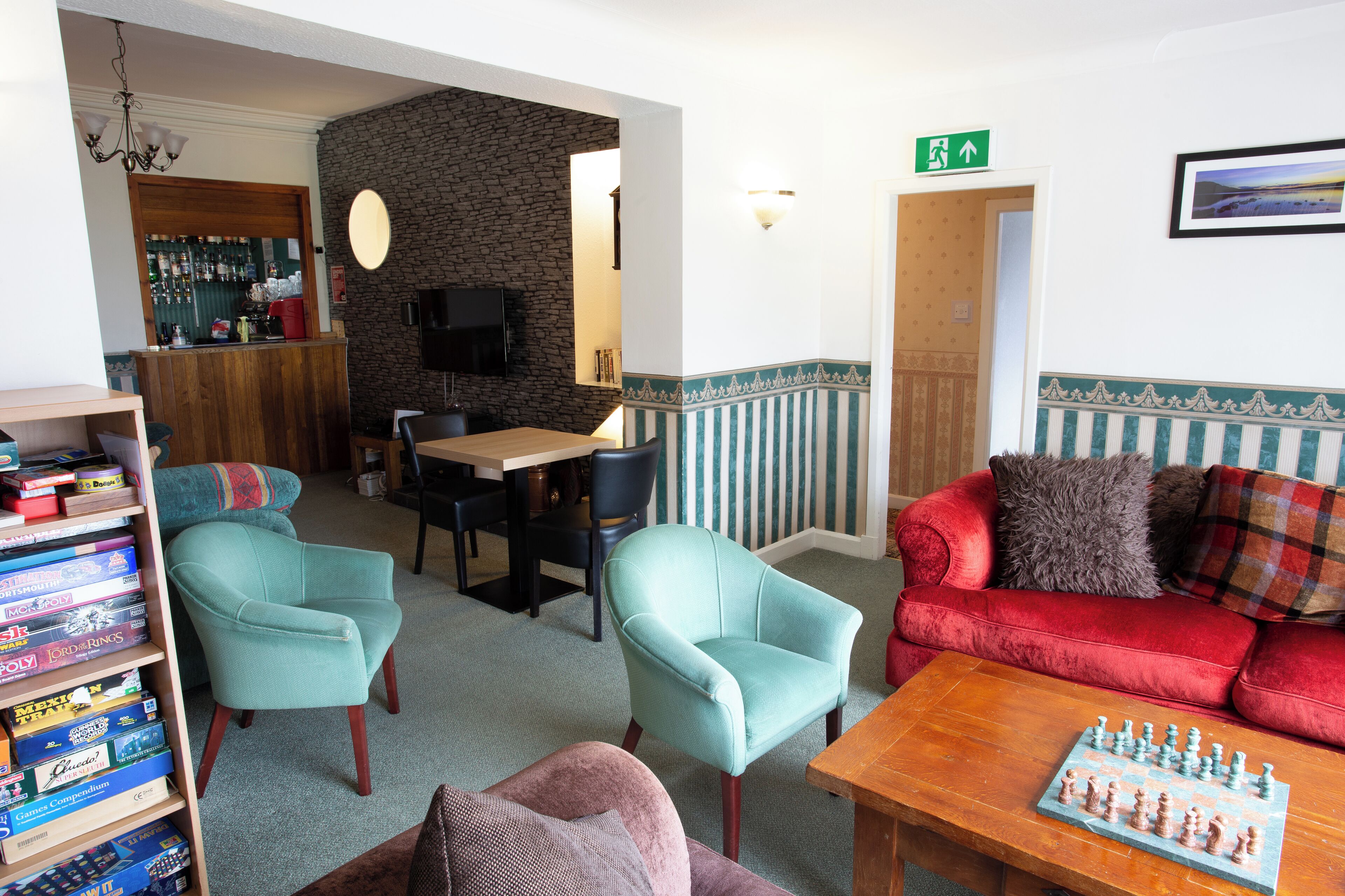 A cozy room with a red sofa, teal armchairs, a wooden coffee table with chess pieces, a bookshelf with board games, a stone wall, and a bar area with a TV.