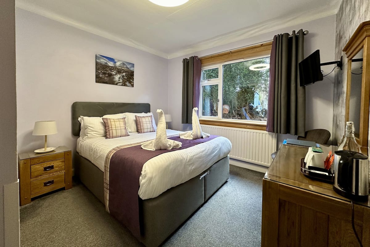 A hotel room with a bed, a bedside table with a lamp, a dresser, a window with curtains, and a painting on the wall.
