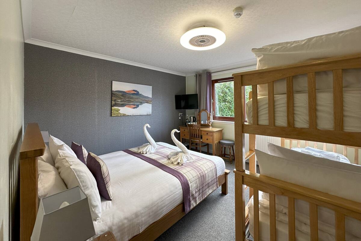 A bedroom with a bed, a desk, a chair, a television, and a window.