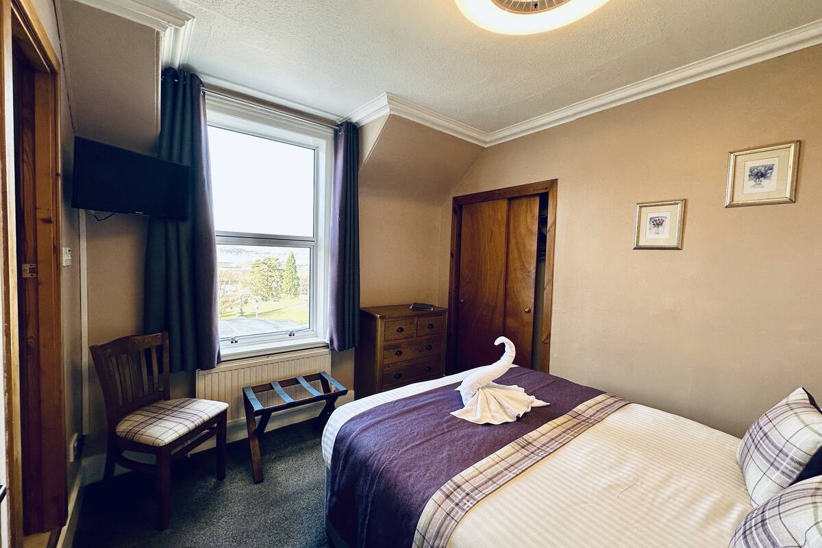 A hotel room with a bed, a chair, a small table, a window with curtains, and a door.