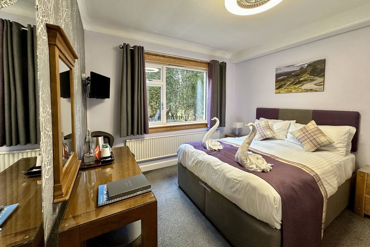 A hotel room with a bed, a desk, a mirror, a window with curtains, a painting on the wall, and a ceiling light.