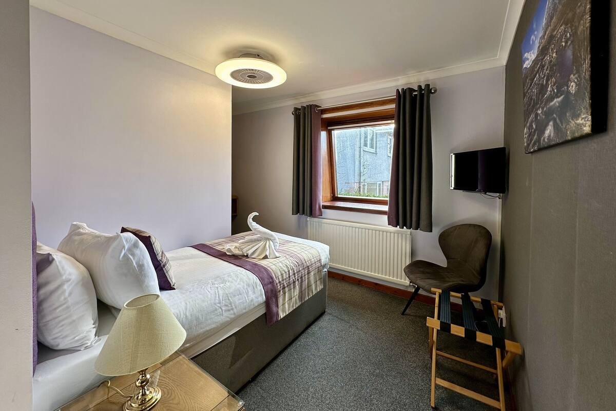 A bedroom with a bed, a desk, a chair, a window with curtains, a radiator, and a flat-screen TV mounted on the wall.