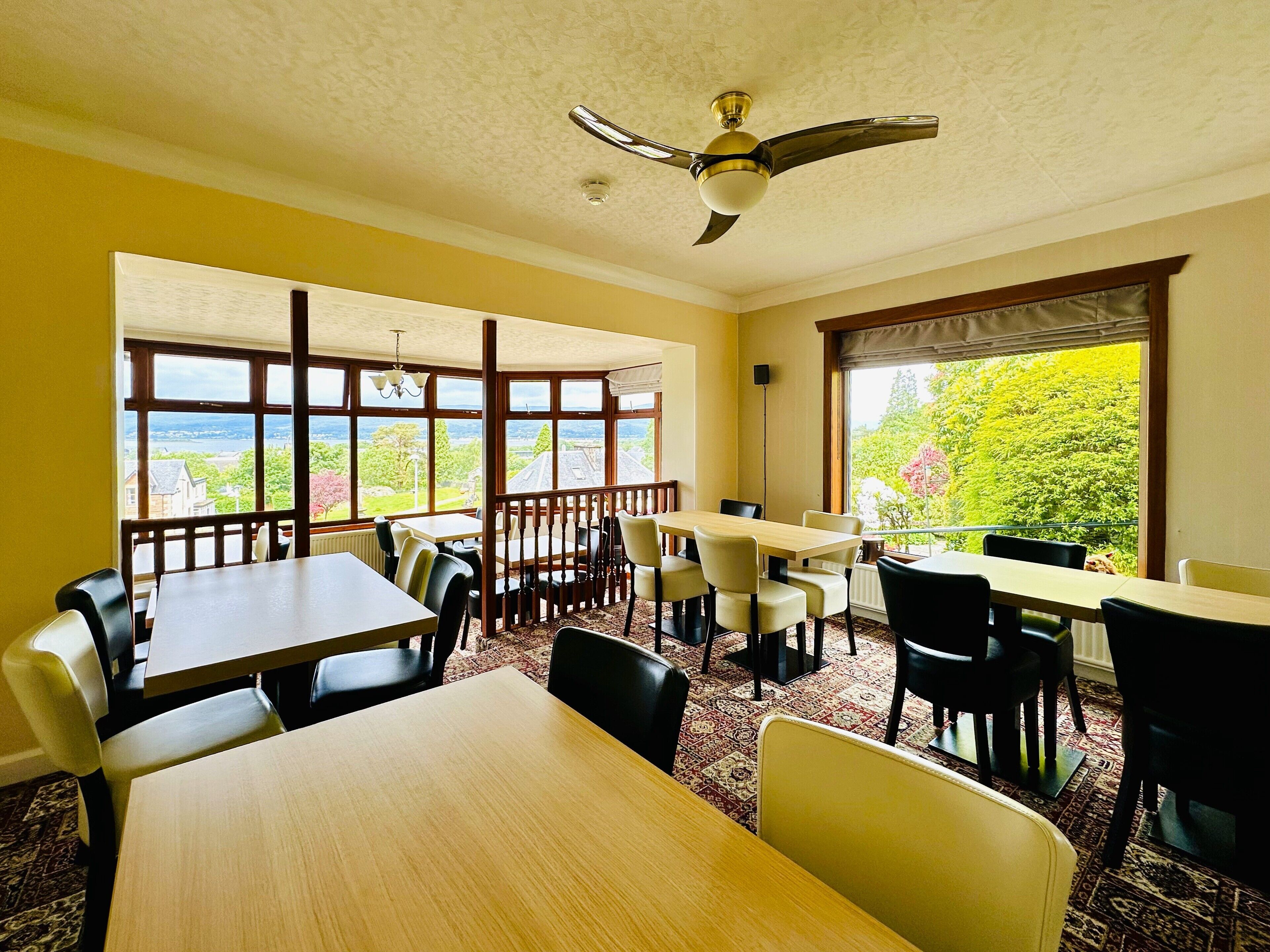 A dining area with tables and chairs, large windows with a view, and a ceiling fan.