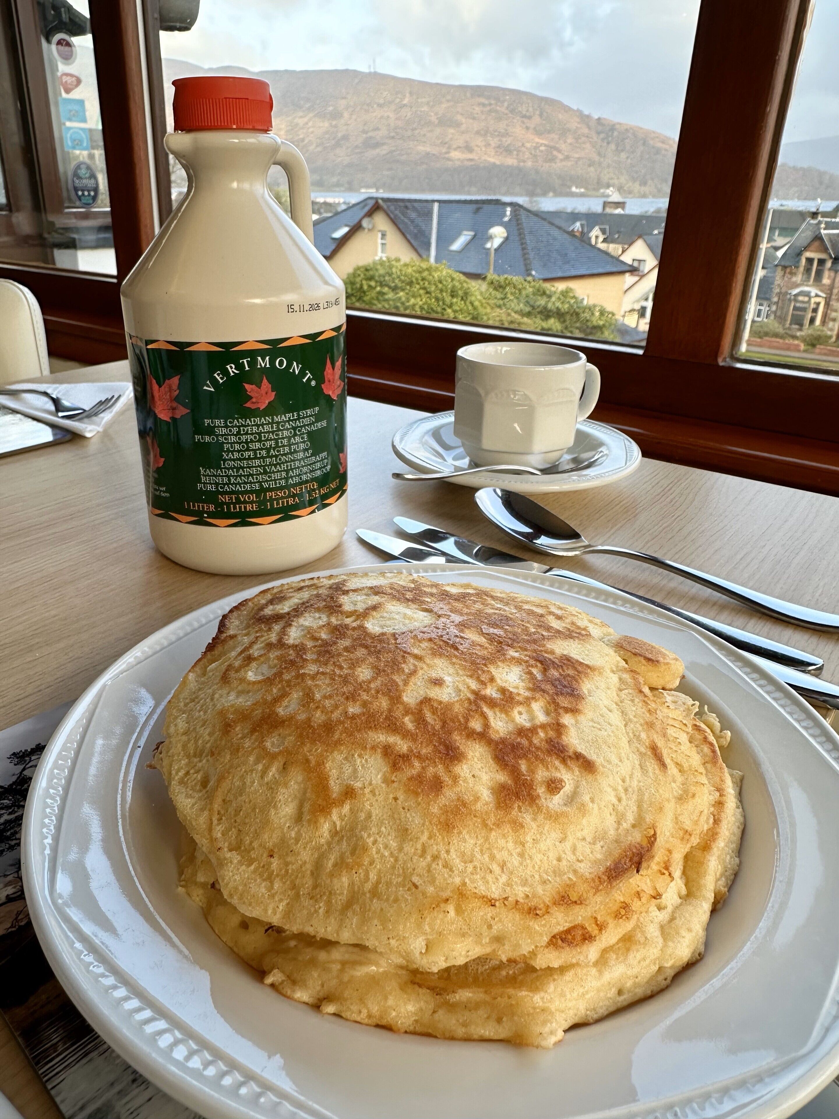 A breakfast setting with pancakes on a plate, a bottle of maple syrup, and a cup on a table by a window with a view of houses and hills.