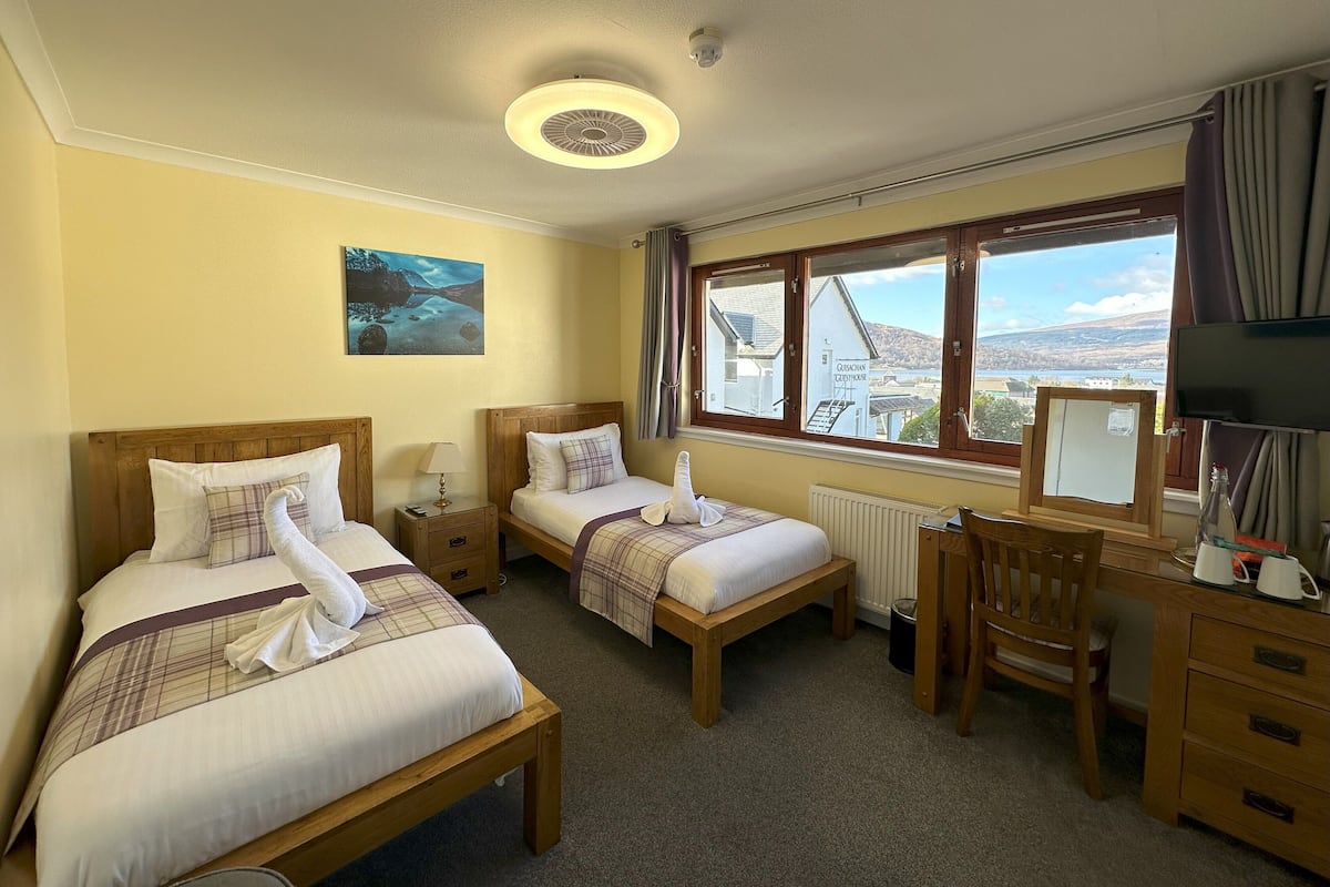 A hotel room with two beds, a desk, a chair, a television, and a window with a view of a building and landscape.