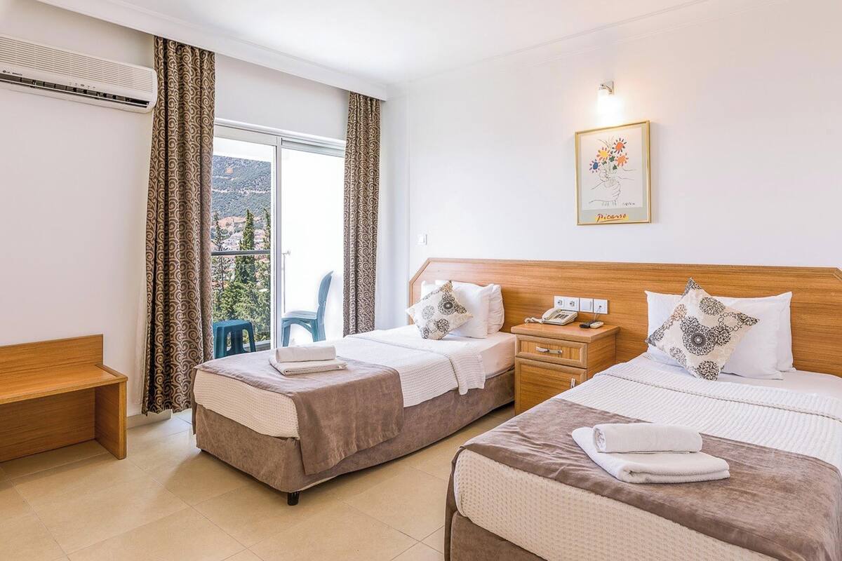 A hotel room with two beds, a wooden headboard, a bedside table with a phone, a framed artwork on the wall, and a view of the outdoors through the window.