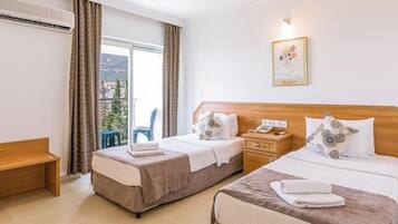 A hotel room with two beds, a wooden headboard, a bedside table with a phone, a framed artwork on the wall, and a view of the outdoors through the window.