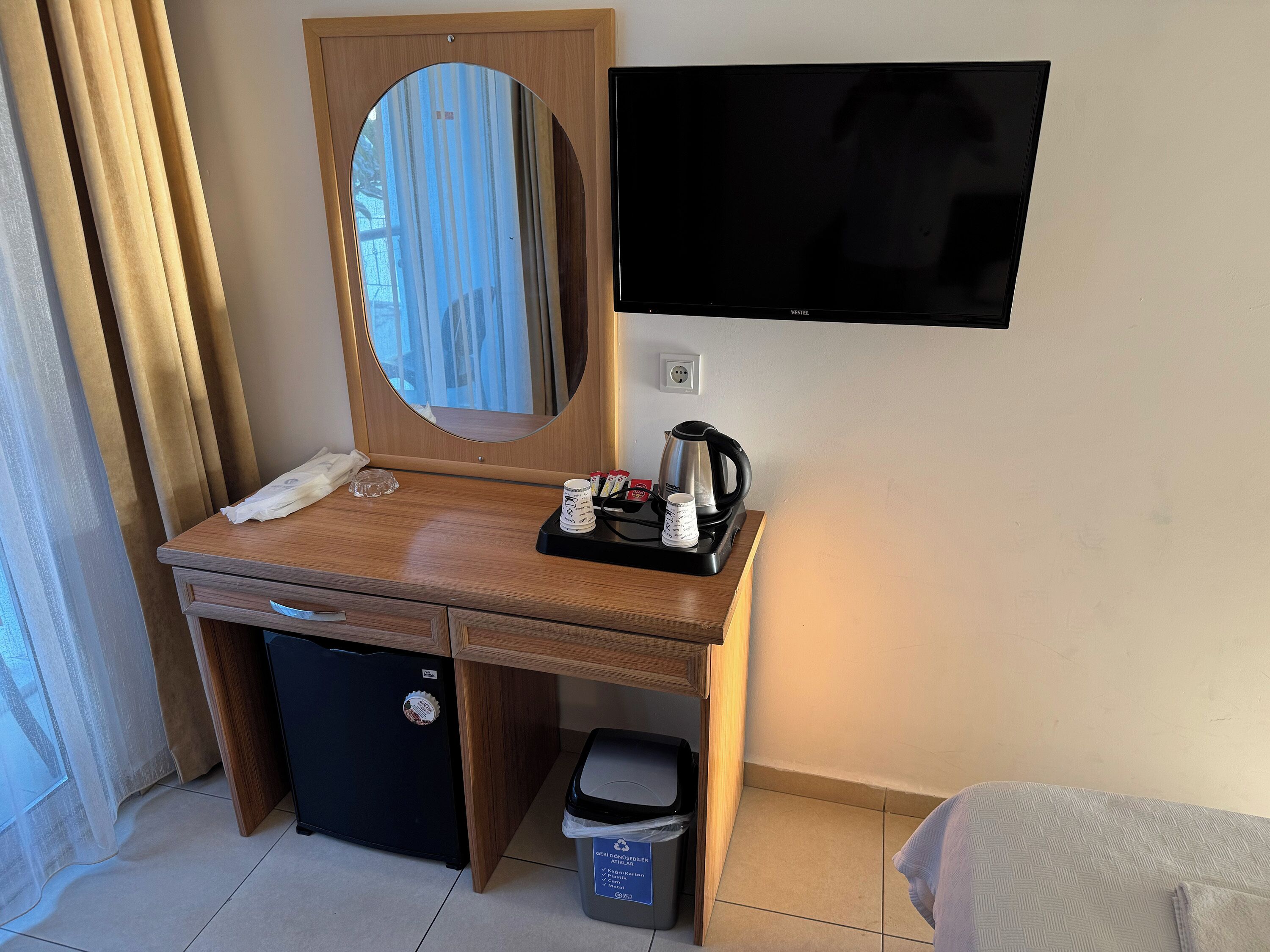 Habitación de hotel compacta con escritorio de madera, espejo, televisor, refrigerador pequeño, cesto de basura y cama con colcha estampada.