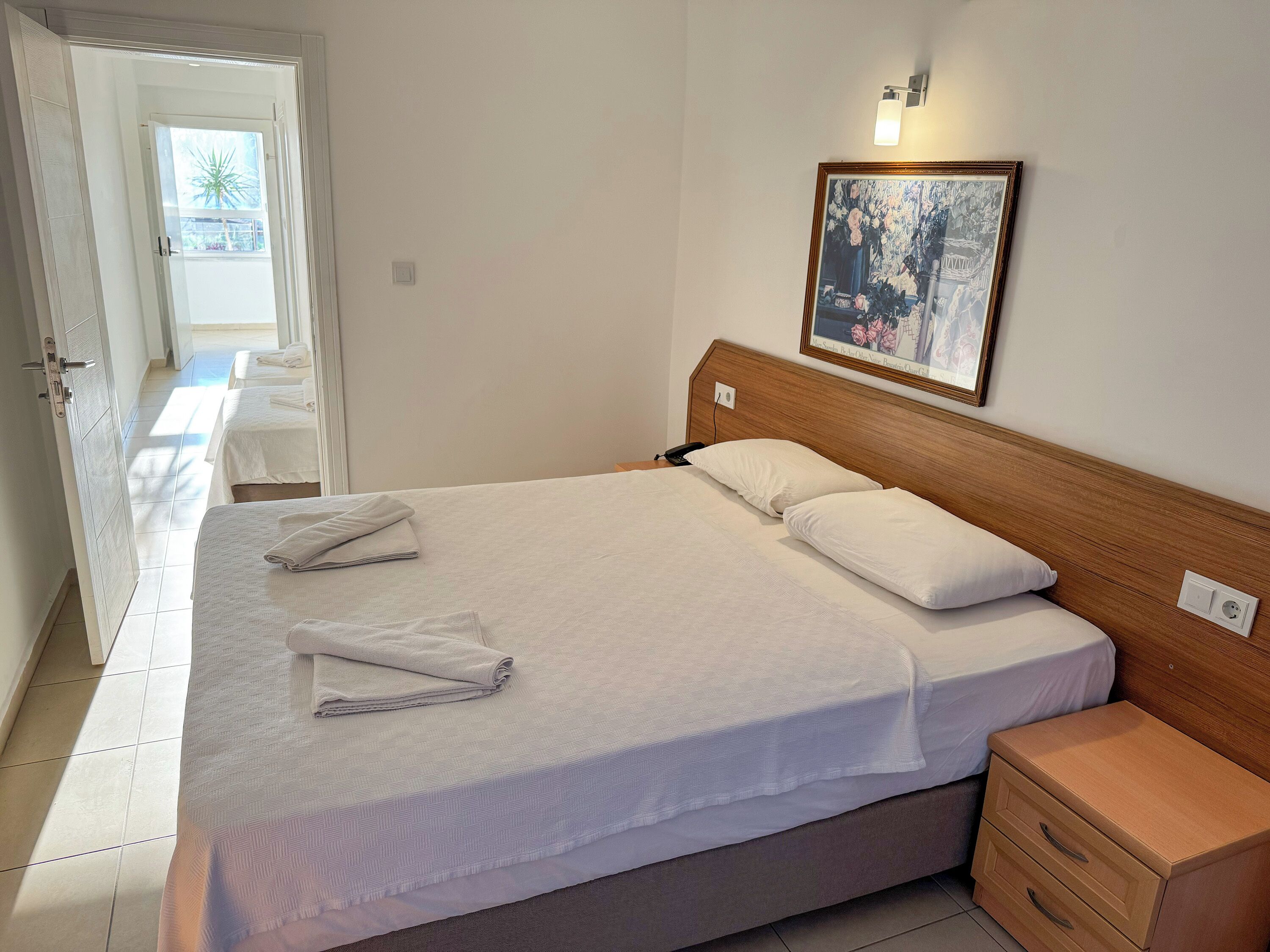 Habitación de hotel con una cama grande, una mesita de noche de madera, un cuadro en la pared y una puerta que da a un balcón.