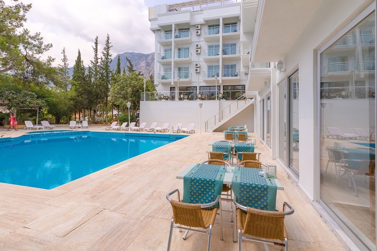 A pool area with lounge chairs and tables, a building with balconies, and a mountainous backdrop.