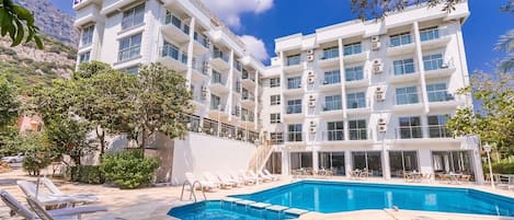 Un hotel con piscina, varios balcones y montañas al fondo.