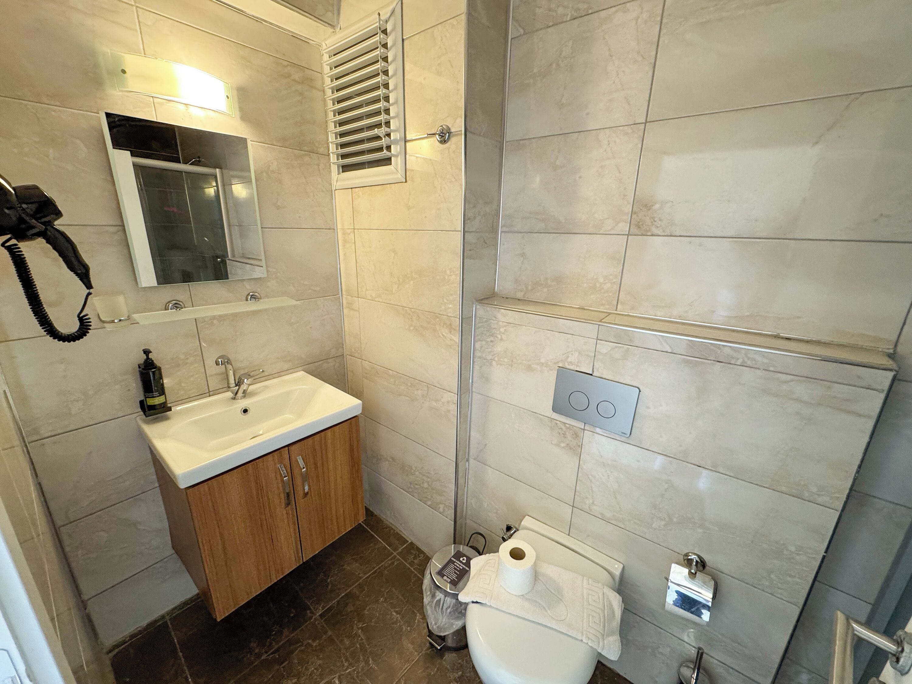 Un baño con lavamanos blanco, armarios de madera, un espejo, inodoro y zona de ducha.