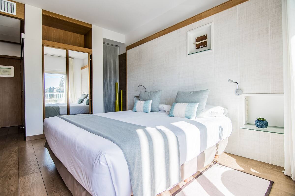 Camera da letto moderna con un letto grande, soffitto in legno, una finestra con vista e un piccolo ripiano con una pianta.