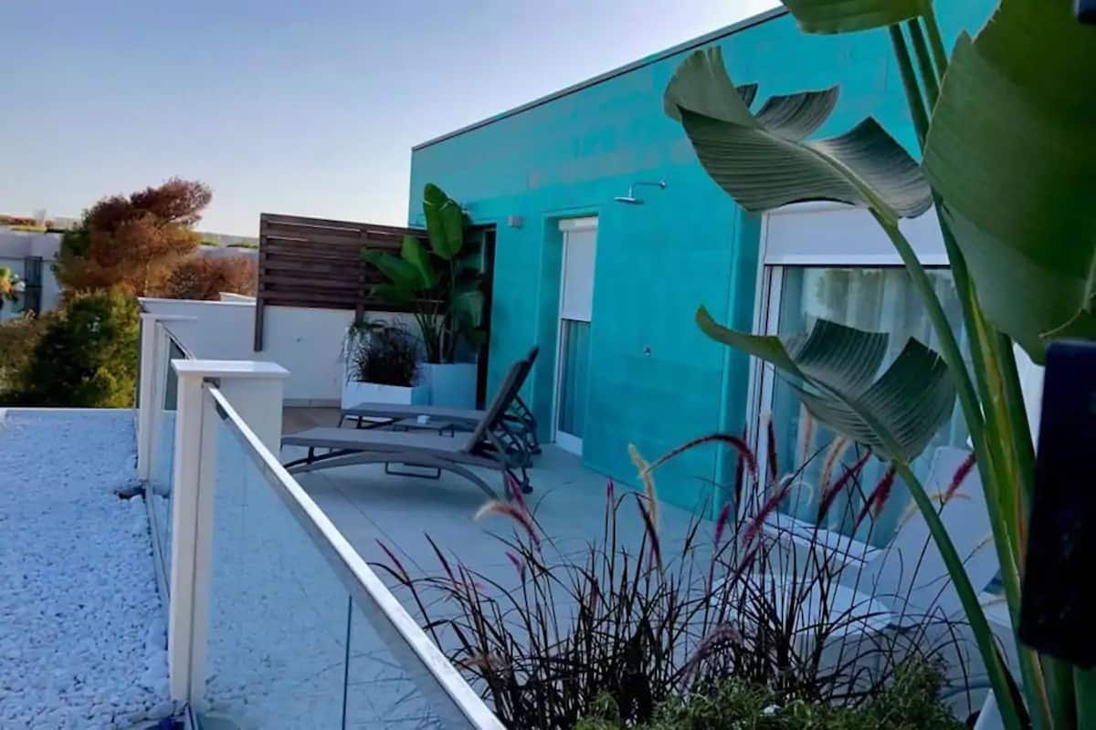 Un edificio moderno con facciata color petrolio, un balcone con ringhiera bianca e un’area salotto con una sedia e un tavolino.