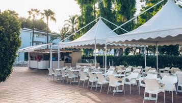 Una terraza con sillas y mesas blancas bajo toldos blancos, una zona de bar y palmeras al fondo.