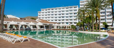 A hotel pool area with sun loungers, umbrellas, and a large building in the background.
