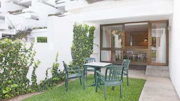 Un patio con mesa y sillas, rodeado de vegetación y un edificio blanco con puerta de cristal.