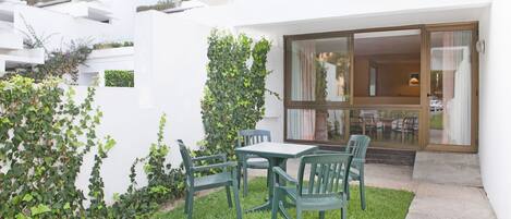 A patio with a table and chairs, surrounded by greenery and a white building with a glass door.
