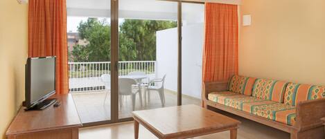 A room with a sofa, a TV, a table, and a balcony with a view of trees.