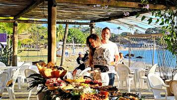 Un área de comedor al aire libre con terraza de madera, sillas de plástico blanco y diversos alimentos en una mesa de buffet.