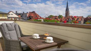 A balcony with wicker furniture and a table set for tea, overlooking a town with traditional buildings and a church spire.
