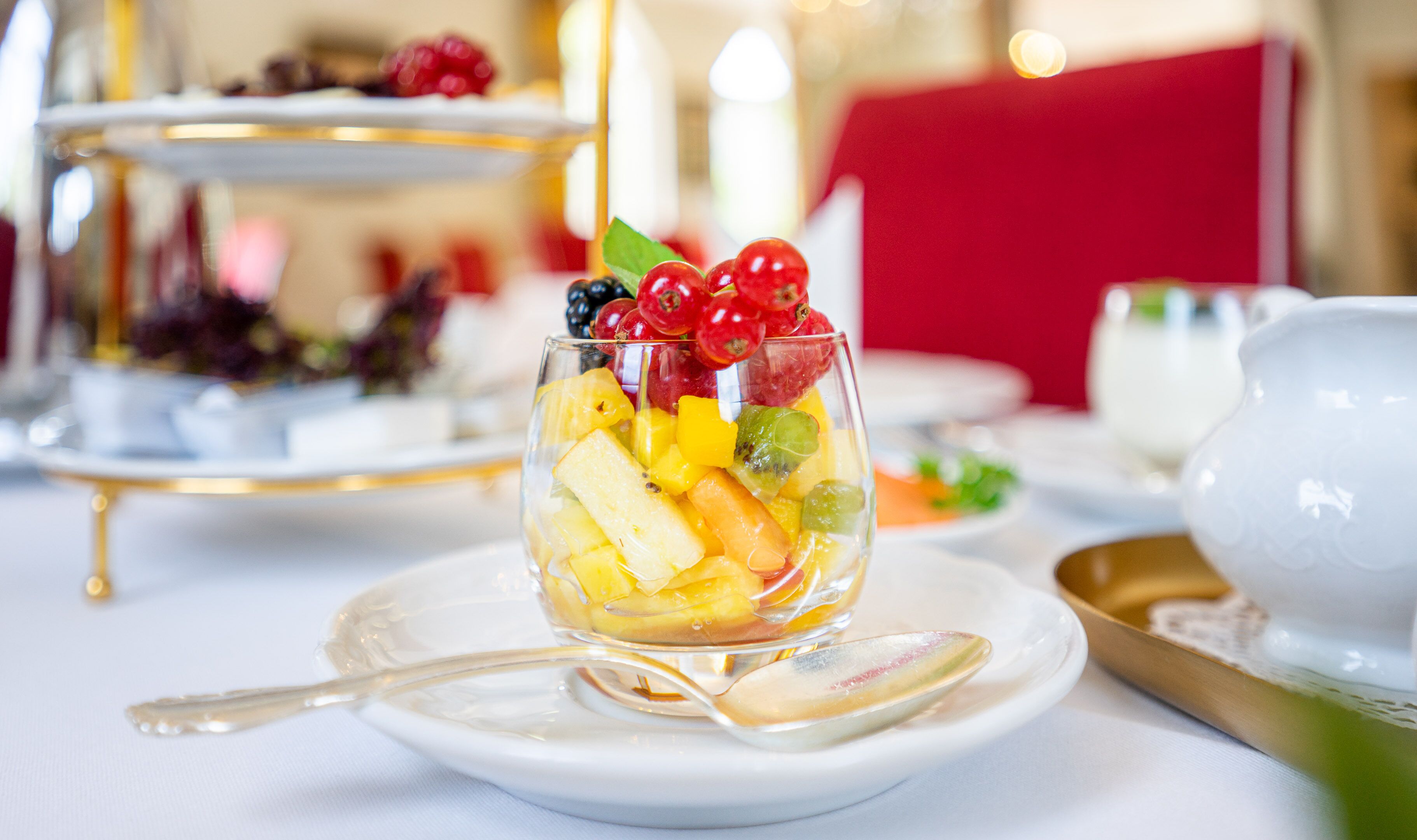 Un tazón de vidrio transparente con fruta variada sobre un plato blanco y una cuchara dorada, sobre un mantel blanco.