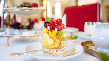Un tazón de vidrio transparente con fruta variada sobre un plato blanco y una cuchara dorada, sobre un mantel blanco.