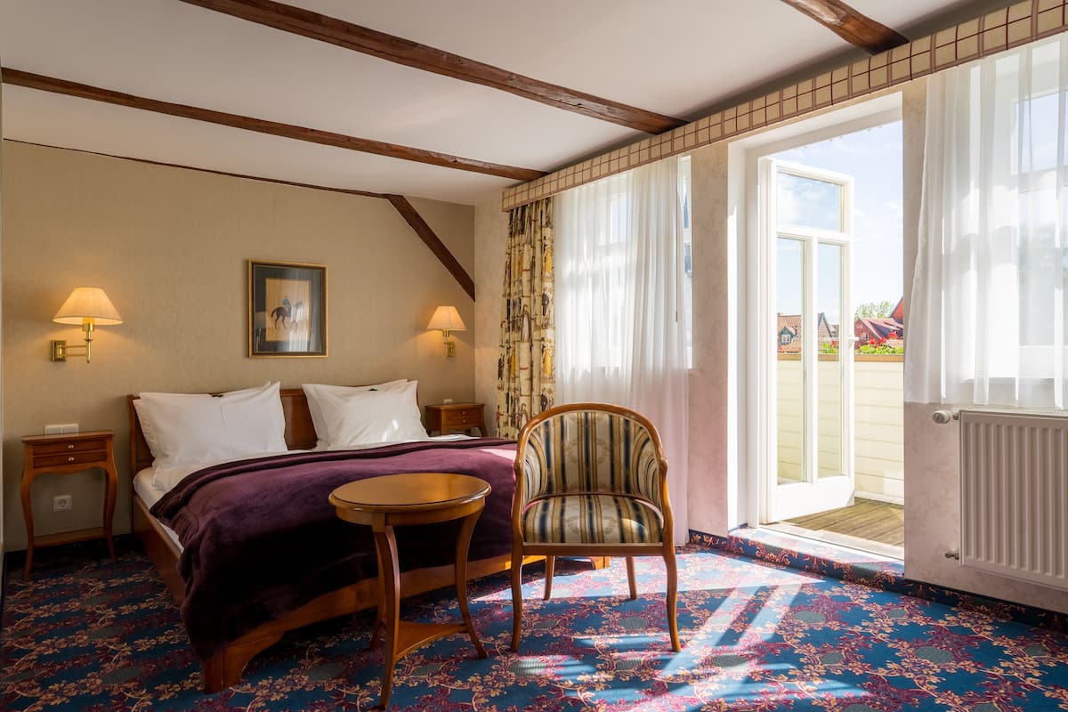 Ein Hotelzimmer mit einem Bett, zwei Nachttischen mit Lampen, einem Sessel, einem kleinen Tisch und Blick ins Freie durch eine Tür.