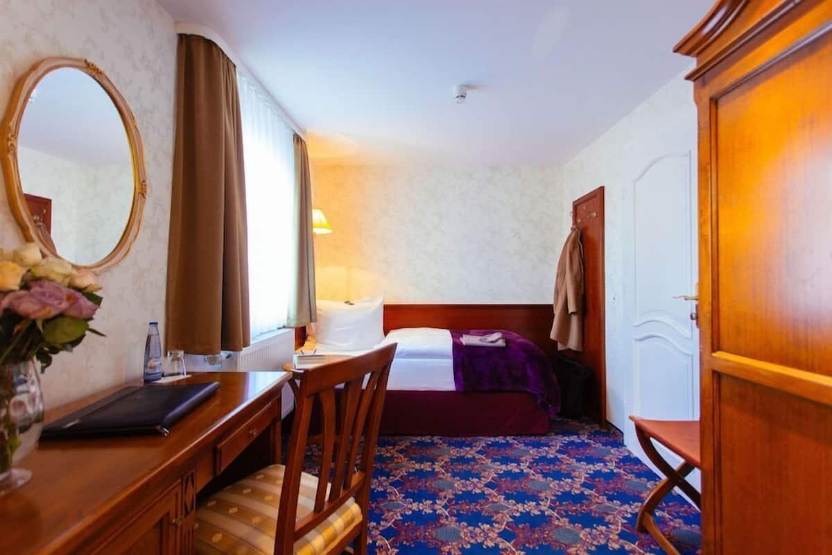 Ein Hotelzimmer mit einem Bett, einem Schreibtisch mit Stuhl, einem Spiegel, einer Kommode und einem Fenster mit Vorhängen.
