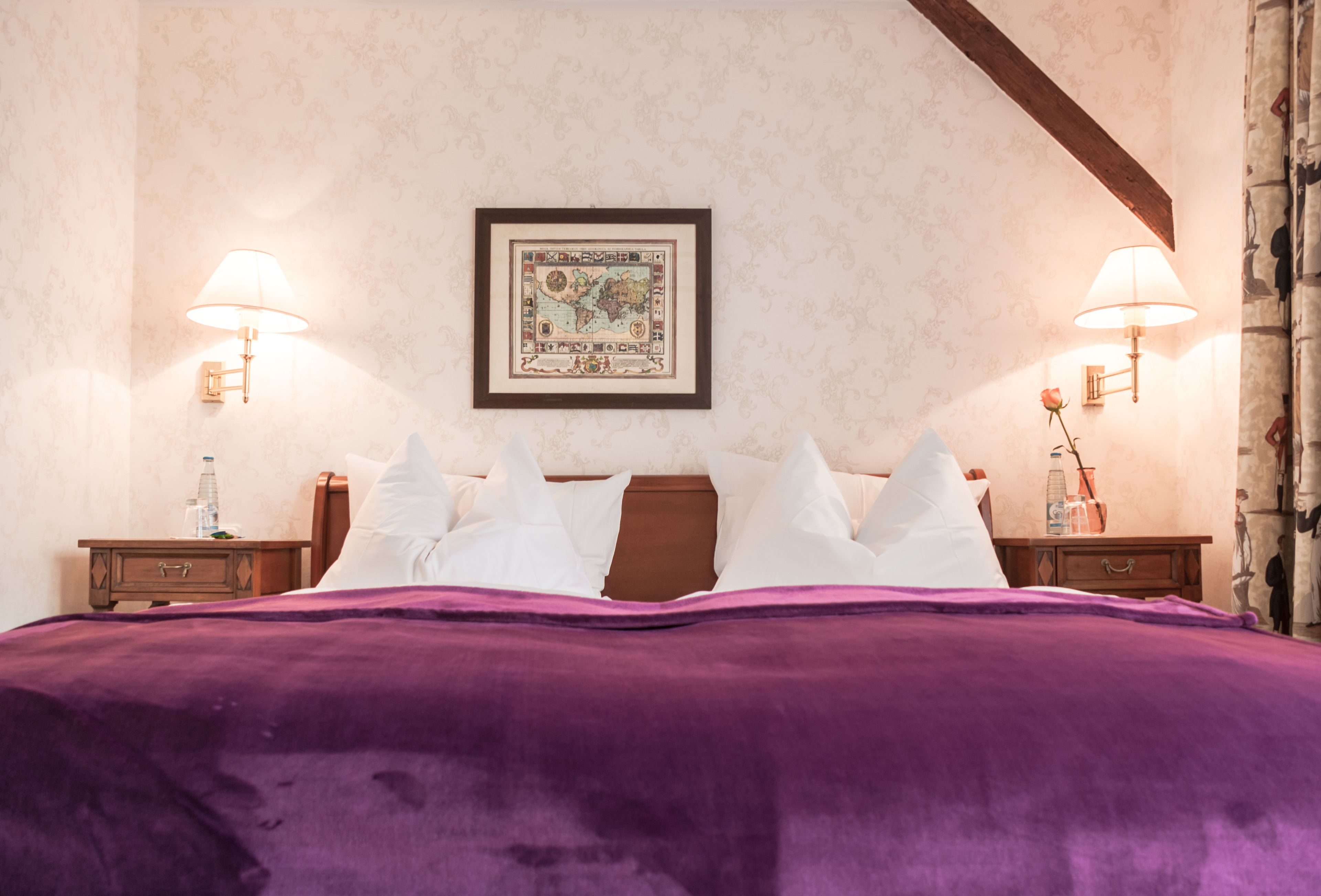 Ein ordentlich bezogenes Bett mit einer violetten Decke, zwei Nachttische mit Lampen und eine gerahmte Karte an der Wand.
