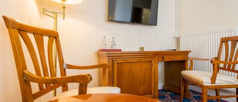 Habitación de hotel con una mesa de comedor de madera, dos sillas de madera, un televisor montado en la pared, una lámpara y un armario con un pequeño ventanal.