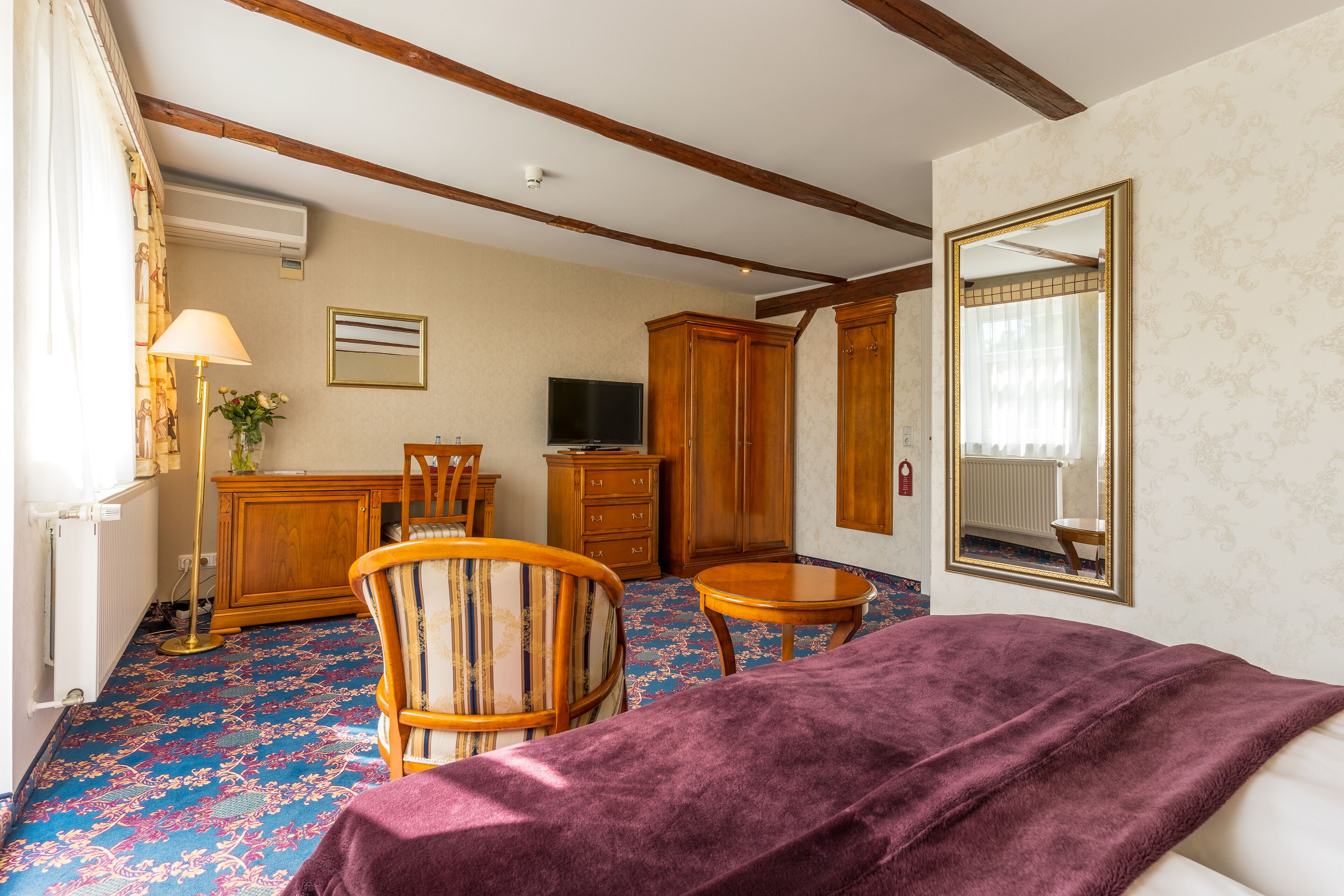 A hotel room with a bed, a chair, a small table, a TV, wooden cabinets, and a mirror.