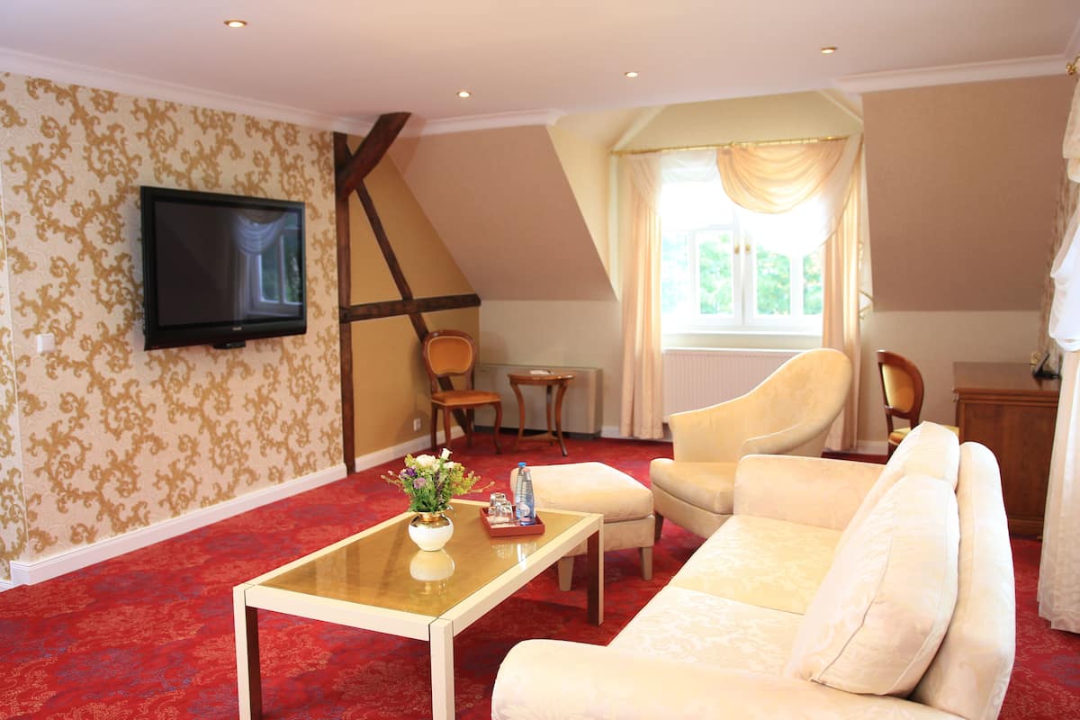 Ein Wohnzimmer mit rotem Teppich, einem beigefarbenen Sofa, einem Holztisch, einem an der Wand montierten Fernseher und einem Fenster mit Vorhängen.