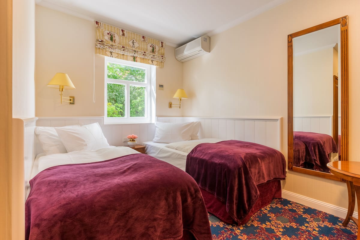 Ein Zimmer mit zwei Betten, jeweils mit violetter Bettdecke, einem Fenster mit Blick auf Grünflächen, Wandlampen und einem großen Spiegel.