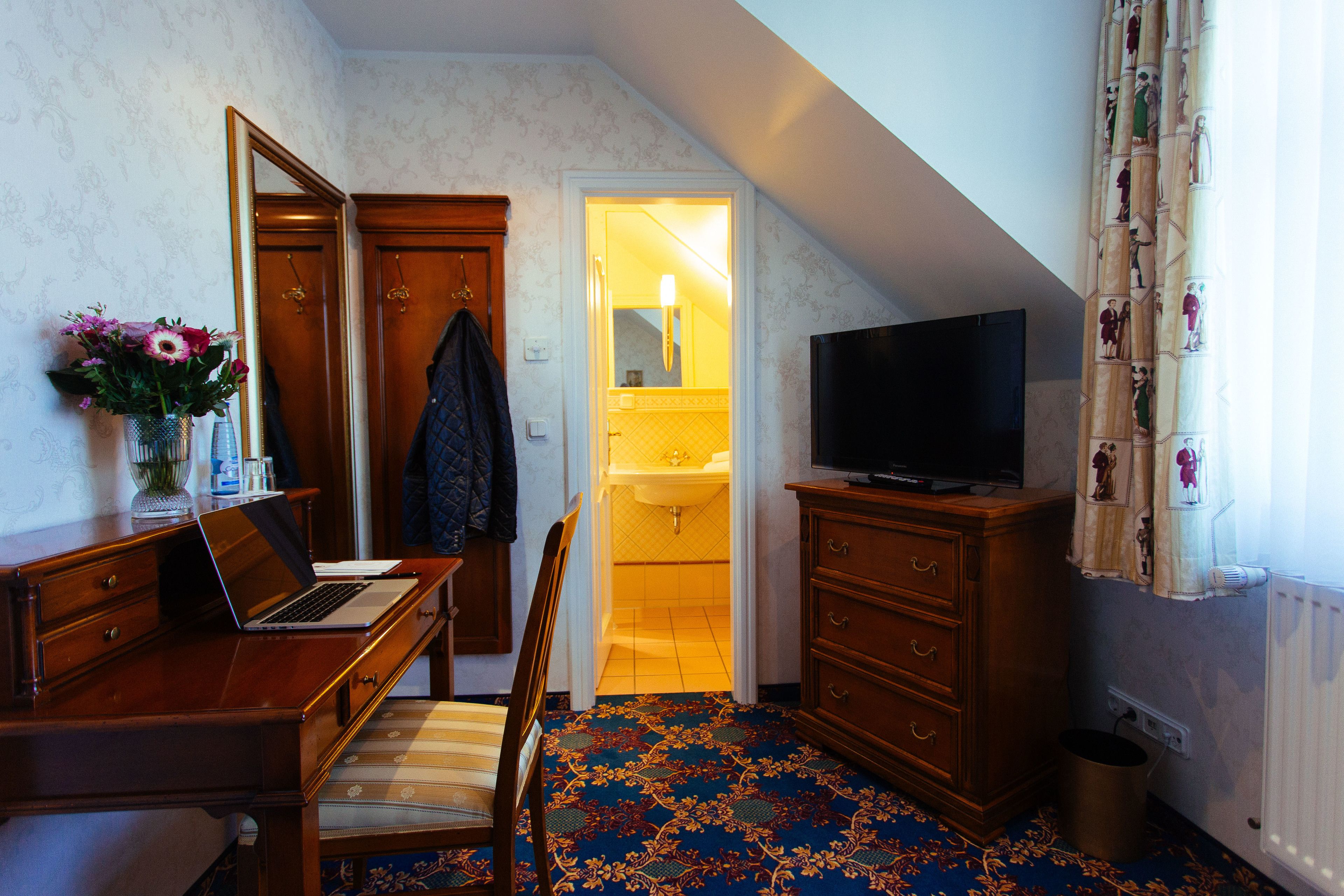 Habitación de hotel con escritorio de madera, una laptop, televisión, silla, un basurero pequeño y un baño visible a través de una puerta abierta.