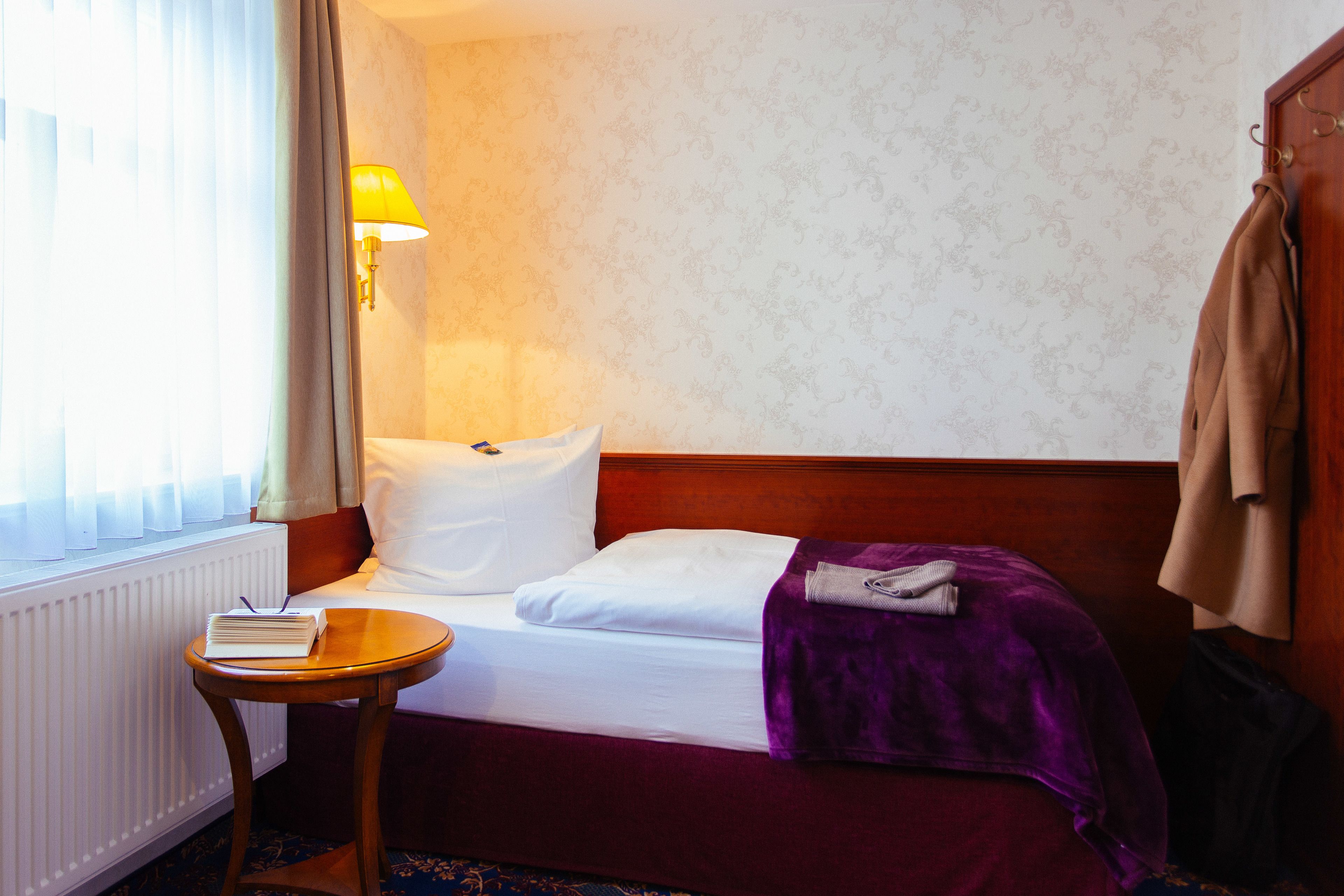 Een hotelkamer met een bed, een nachtkastje met een boek, een lamp en een kapstok.