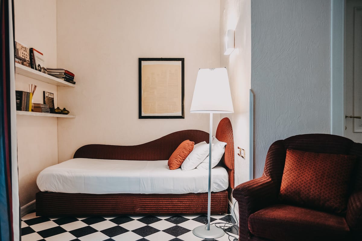 A neatly arranged hotel room with a bed, a chair, a lamp, and a shelf with books and a framed picture.
