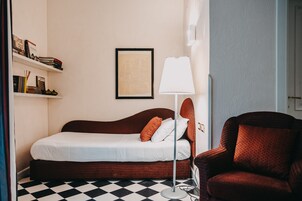 Camera d'albergo ordinata con un letto, una sedia, una lampada e una mensola con libri e una cornice.