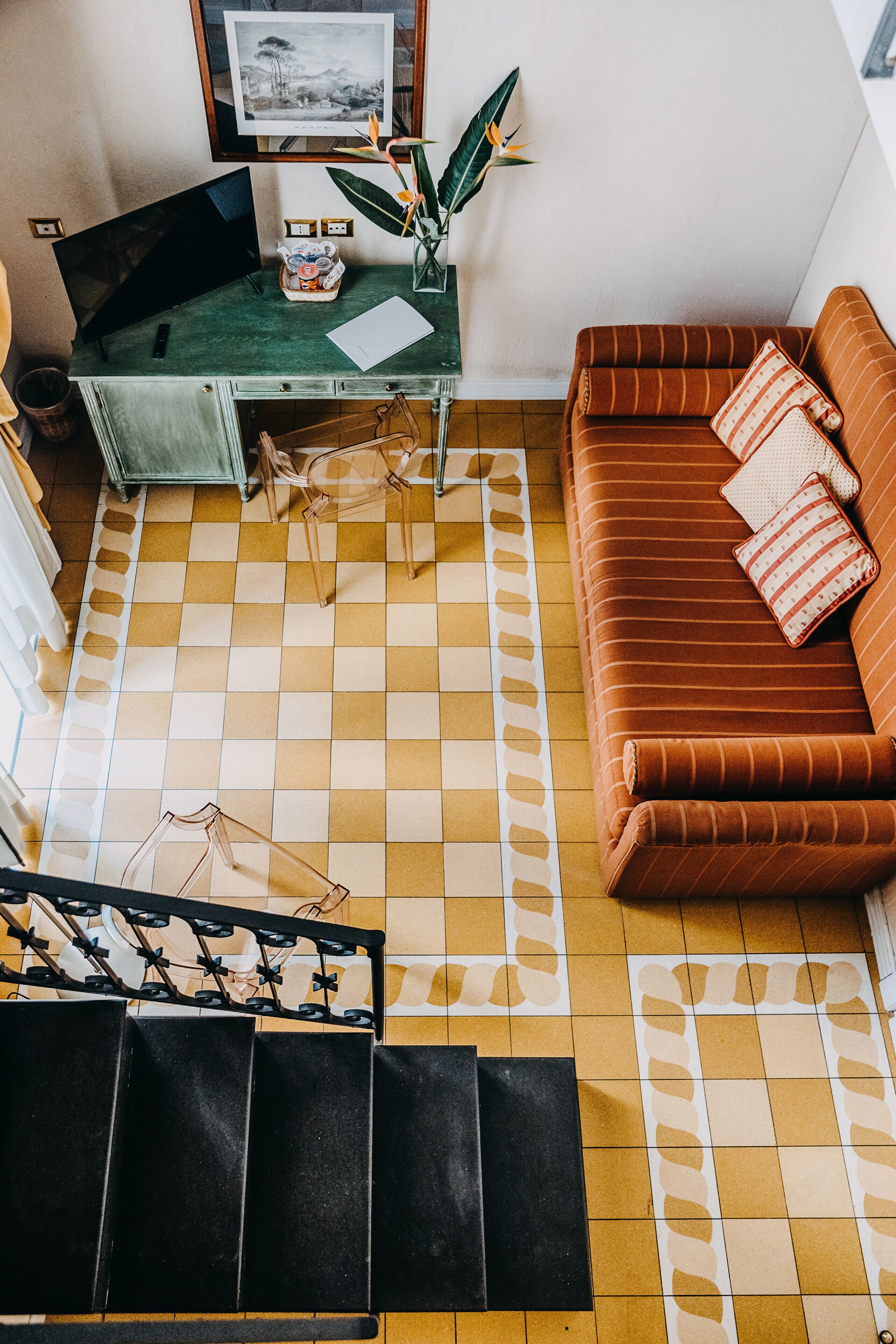 Un salon avec un sol à carreaux, un canapé marron agrémenté de coussins à rayures, un bureau vert avec un ordinateur et une plante.