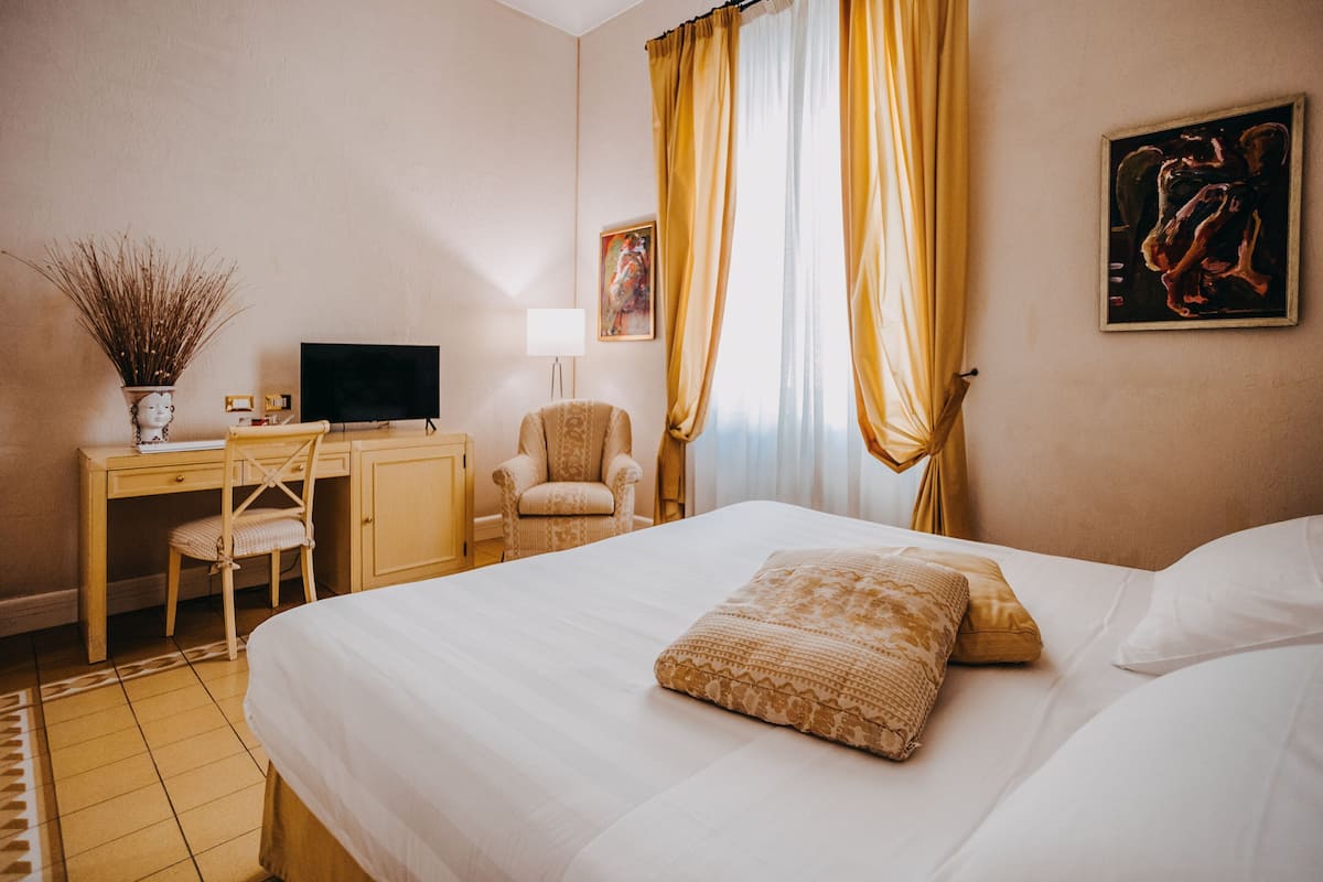 A hotel room with a bed, a desk with a computer, a chair, and a painting on the wall.