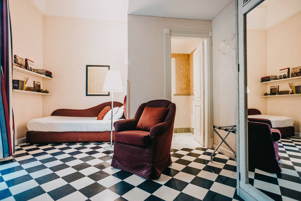 A hotel room with a checkered floor, a red armchair, a sofa, and a bed.