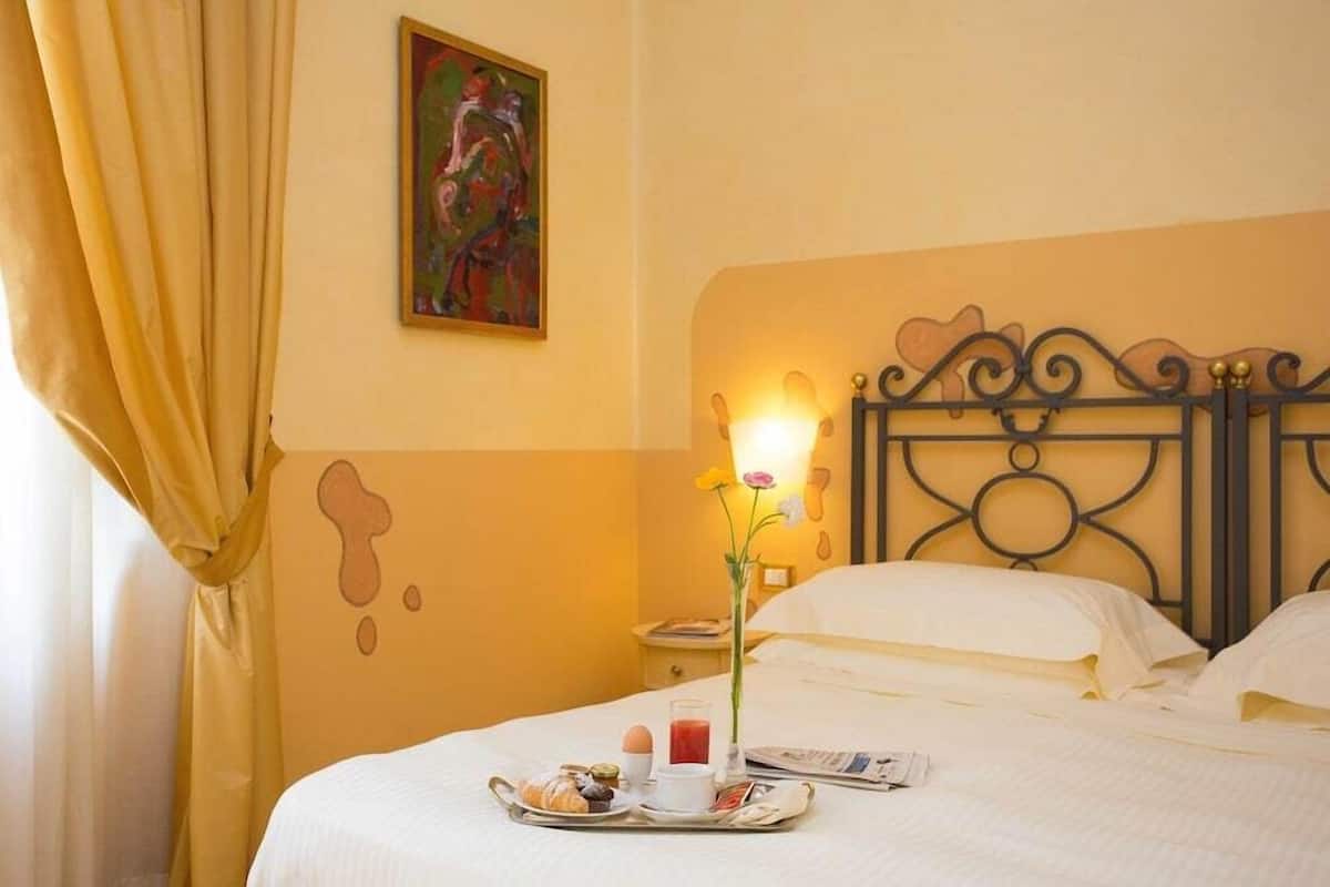 A hotel room with a bed, a bedside table with a lamp, a painting on the wall, and a breakfast tray on the bed.