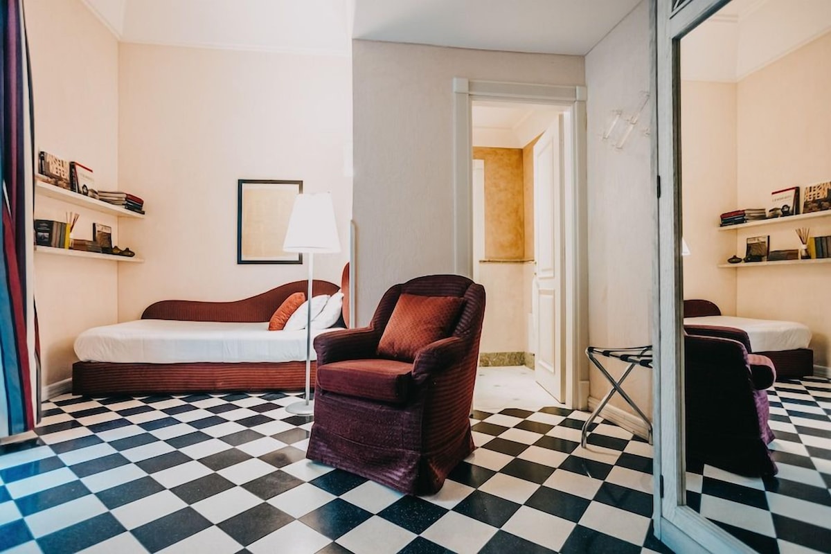 A hotel room with a checkered floor, a bed, a chair, and a mirror.