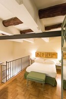Een slaapkamer met een houten plafond, een bed met groene beddengoed, een bankje en een balustrade langs het balkon.